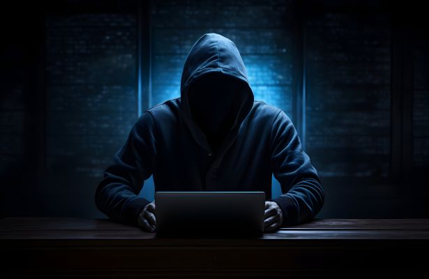 man in a hoody, with his face covered, writing on a laptop.